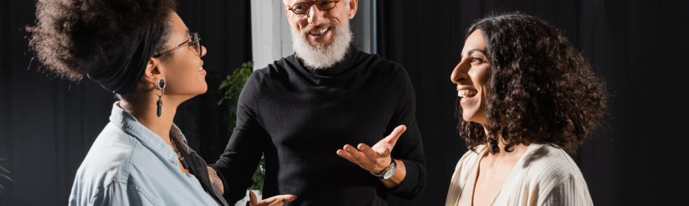 positive grey haired producer pointing with hand while talking to cheerful interracial actresses in theater